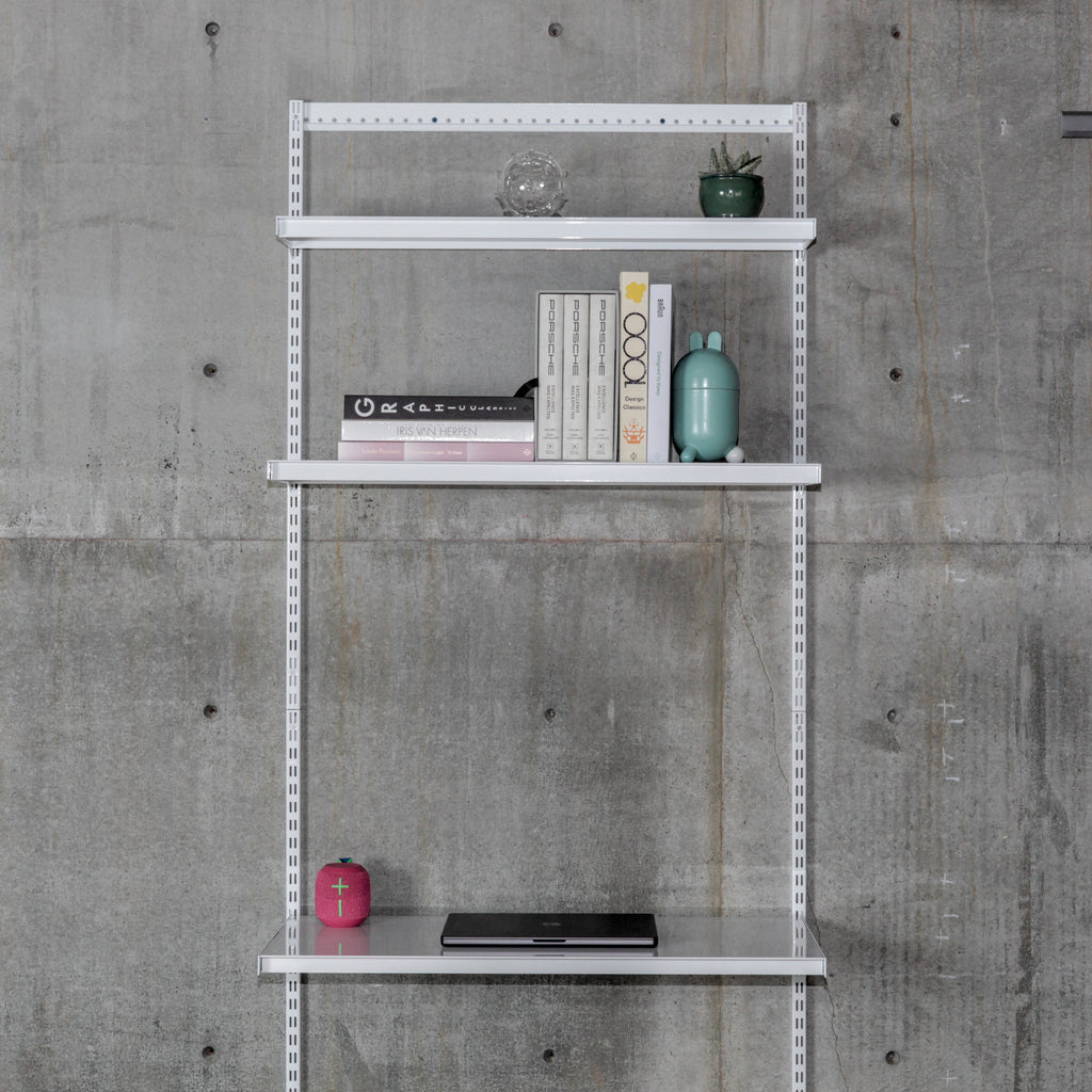 Modular wall-mounted steel shelving system with three white shelves on vertical standards against concrete wall