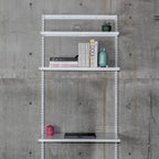 Modular wall-mounted steel shelving system with three white shelves on vertical standards against concrete wall