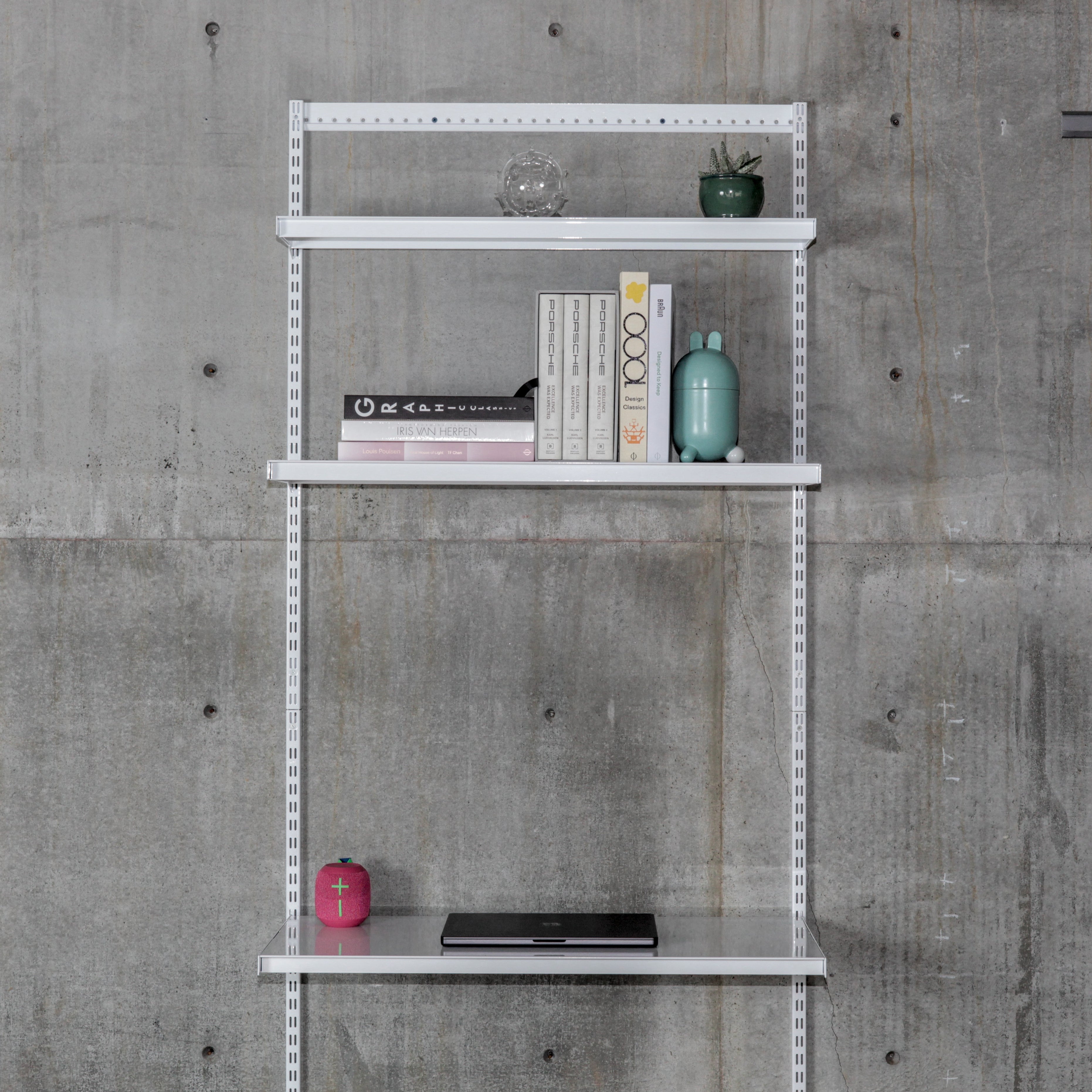 Modular wall-mounted steel shelving system with three white shelves on vertical standards against concrete wall