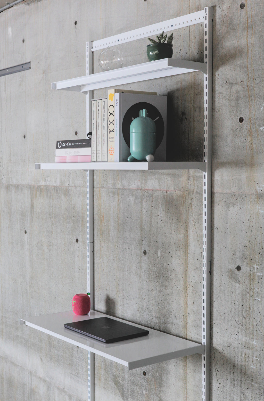 Modular wall-mounted steel shelving system with three white shelves on vertical standards against concrete wall