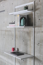 Modular wall-mounted steel shelving system with three white shelves on vertical standards against concrete wall
