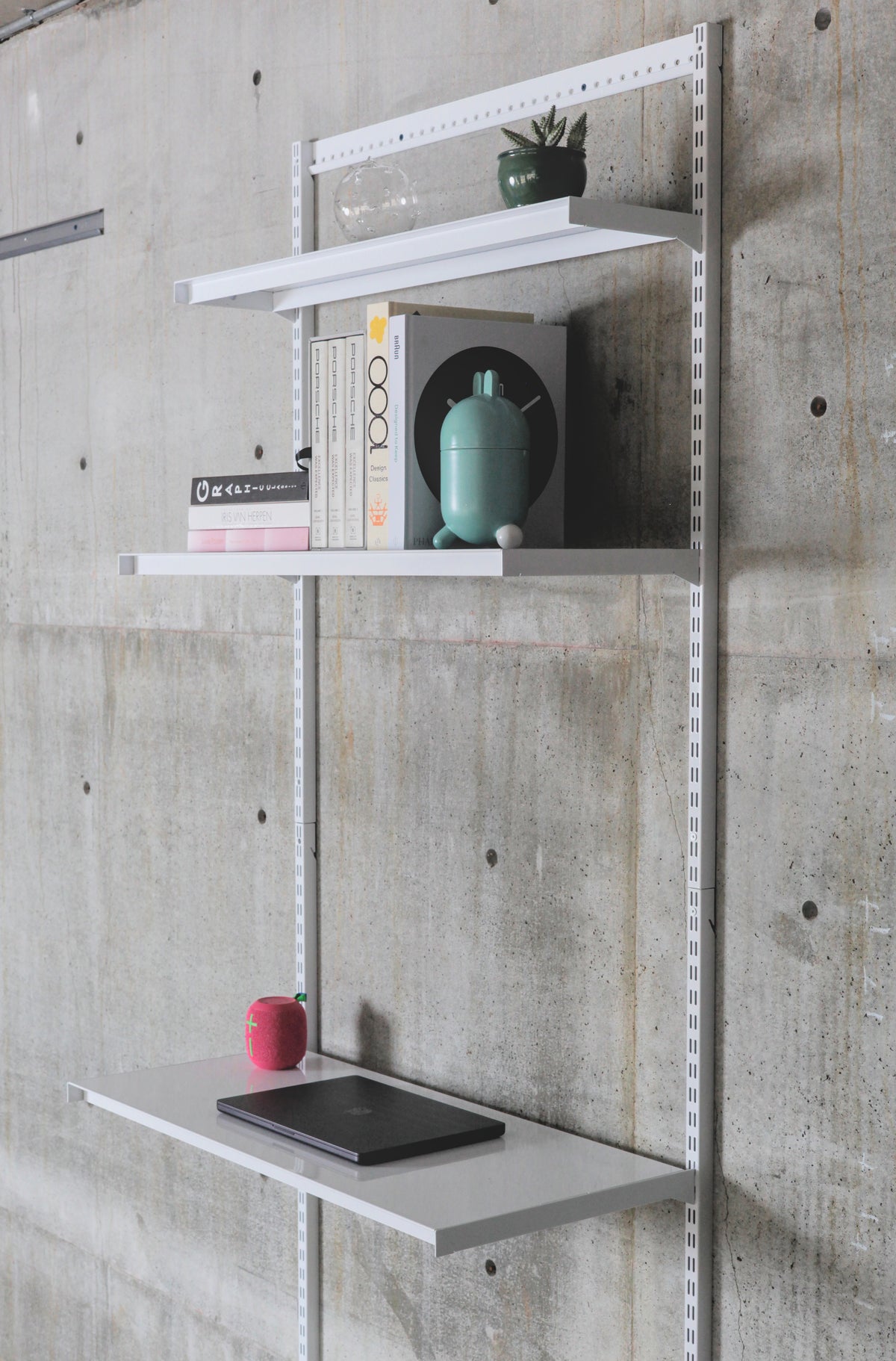 Modular wall-mounted steel shelving system with three white shelves on vertical standards against concrete wall