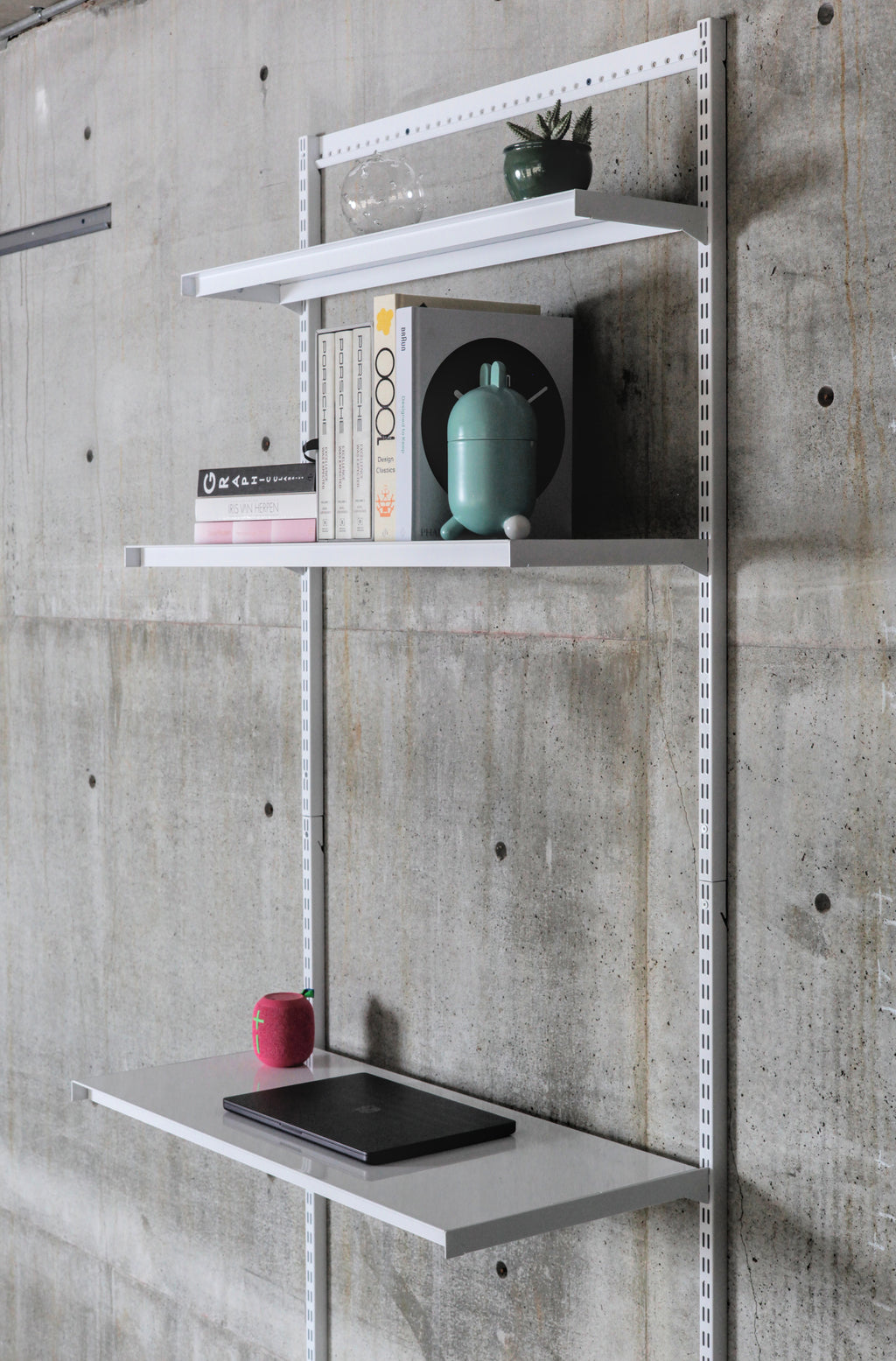 Modular wall-mounted steel shelving system with three white shelves on vertical standards against concrete wall