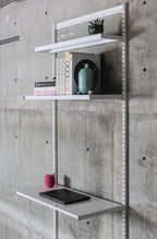 Modular wall-mounted steel shelving system with three white shelves on vertical standards against concrete wall