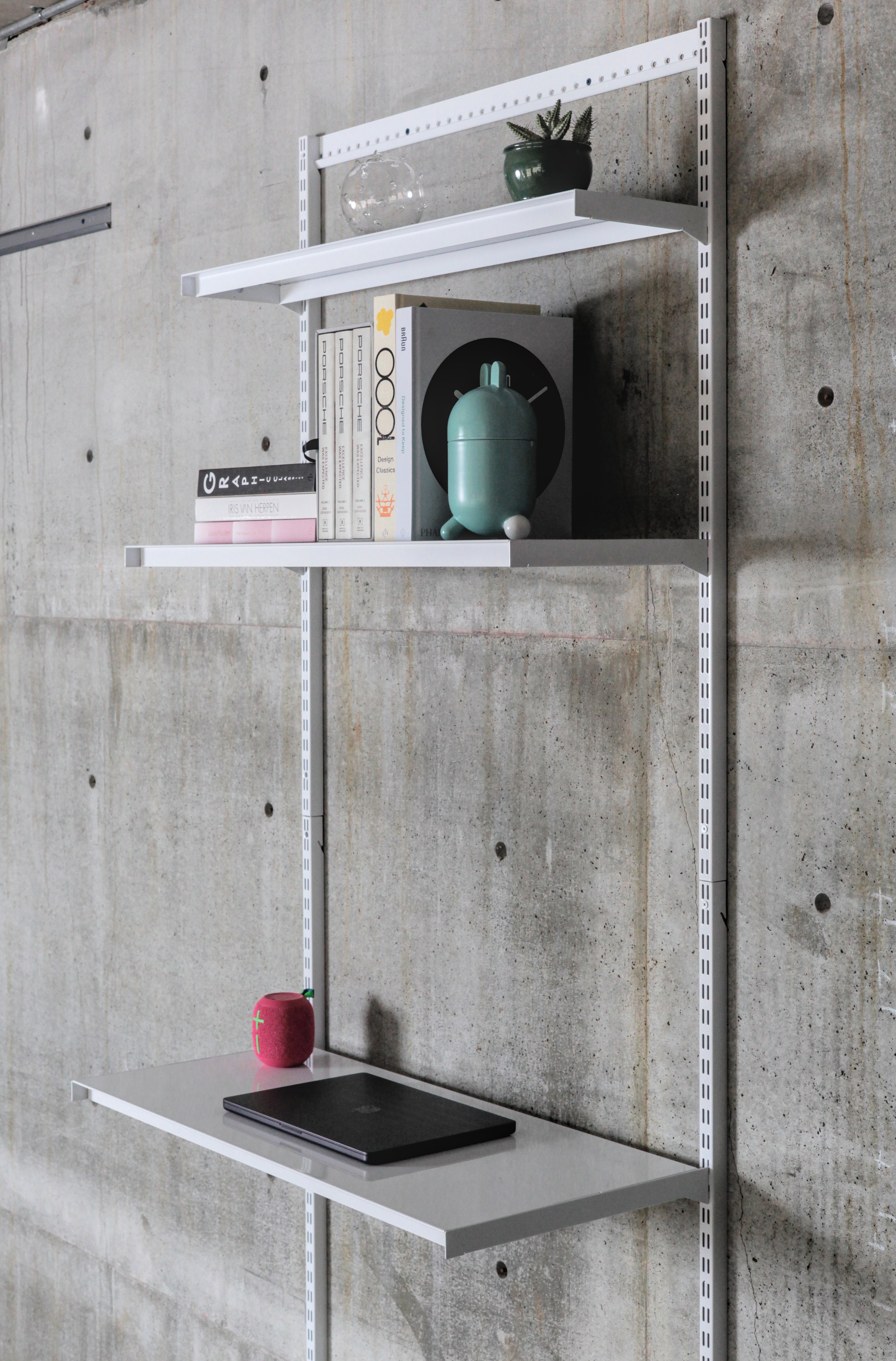 Modular wall-mounted steel shelving system with three white shelves on vertical standards against concrete wall