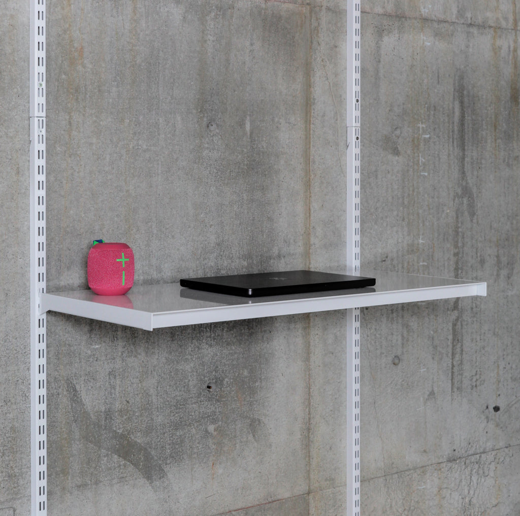 A single adjustable metal modular shelf with laptop and bluetooth speaker mounted to standards on a grey concrete wall