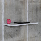 A single adjustable metal modular shelf with laptop and bluetooth speaker mounted to standards on a grey concrete wall