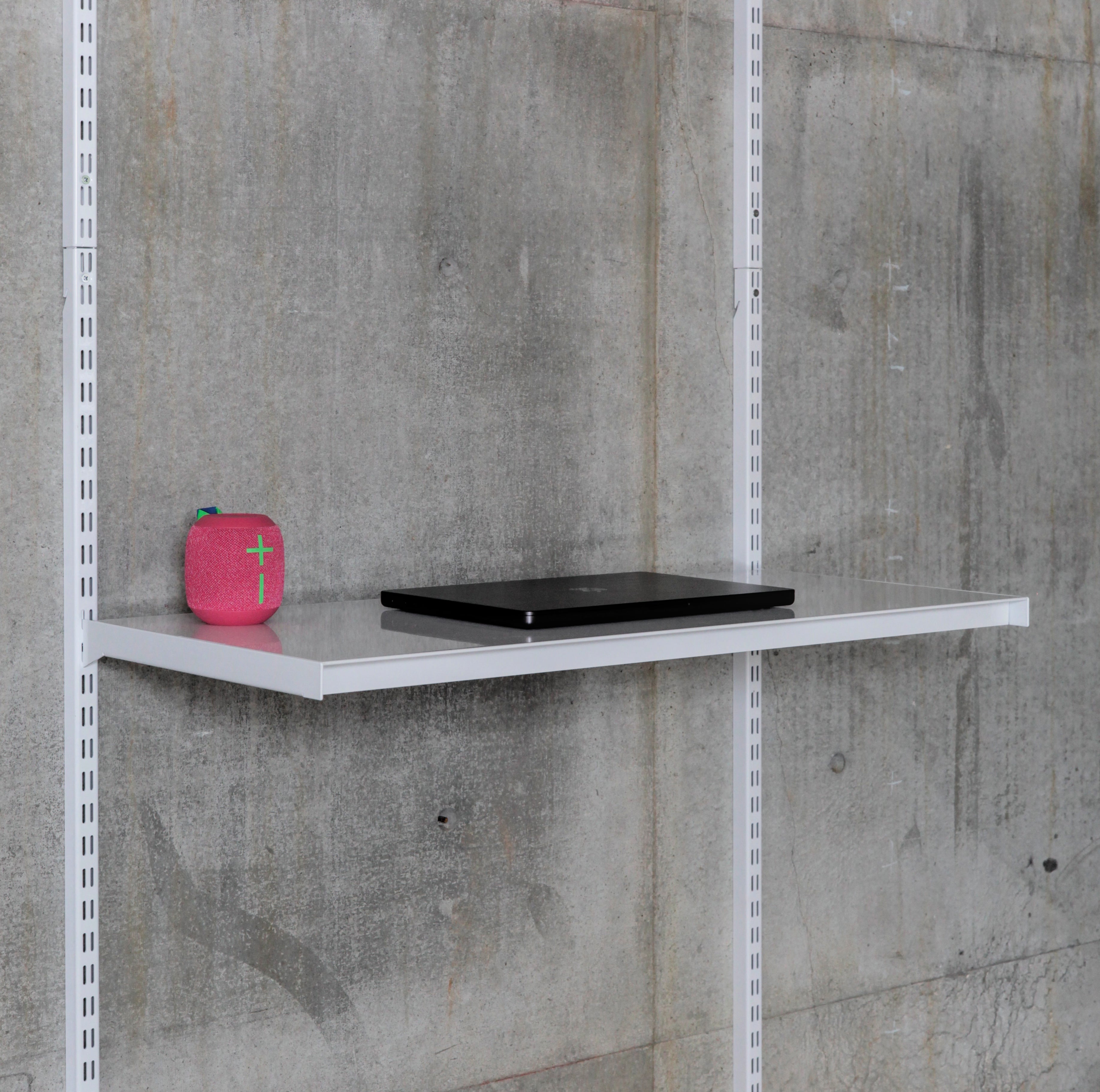 A single adjustable metal modular shelf with laptop and bluetooth speaker mounted to standards on a grey concrete wall